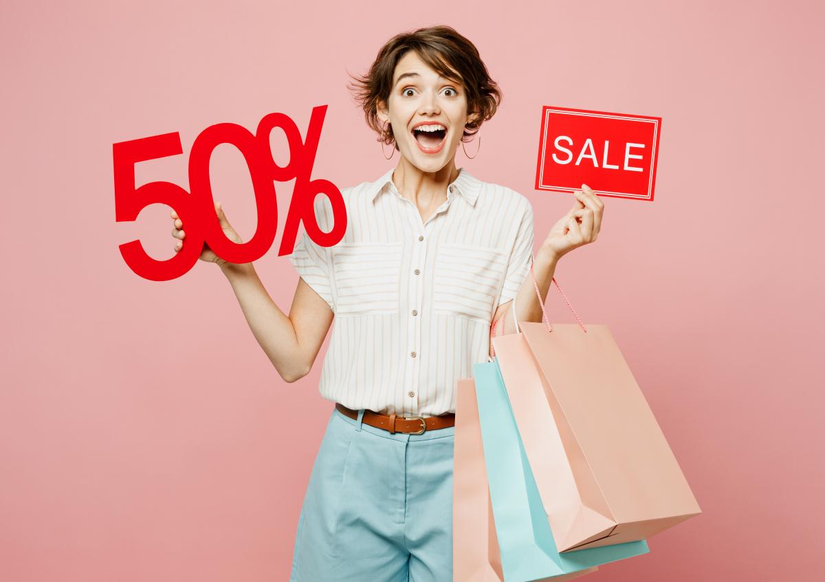A cheerful young woman holding several shopping bags in one hand and red sale signs showing "50%" and "SALE" in the other, expressing excitement and enthusiasm.