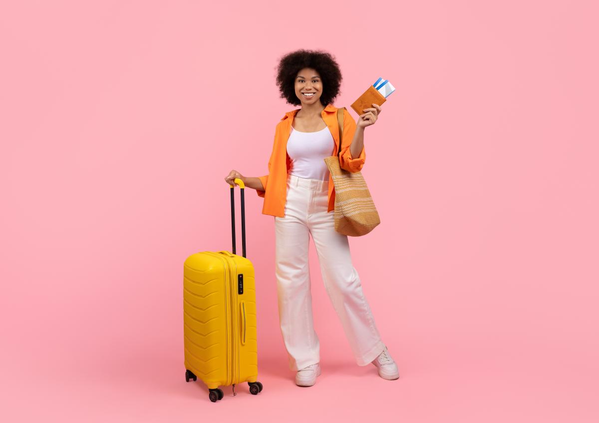 A woman stands smiling confidently while holding a suitcase with one hand and a wallet with travel tickets in the other hand, ready for a trip.