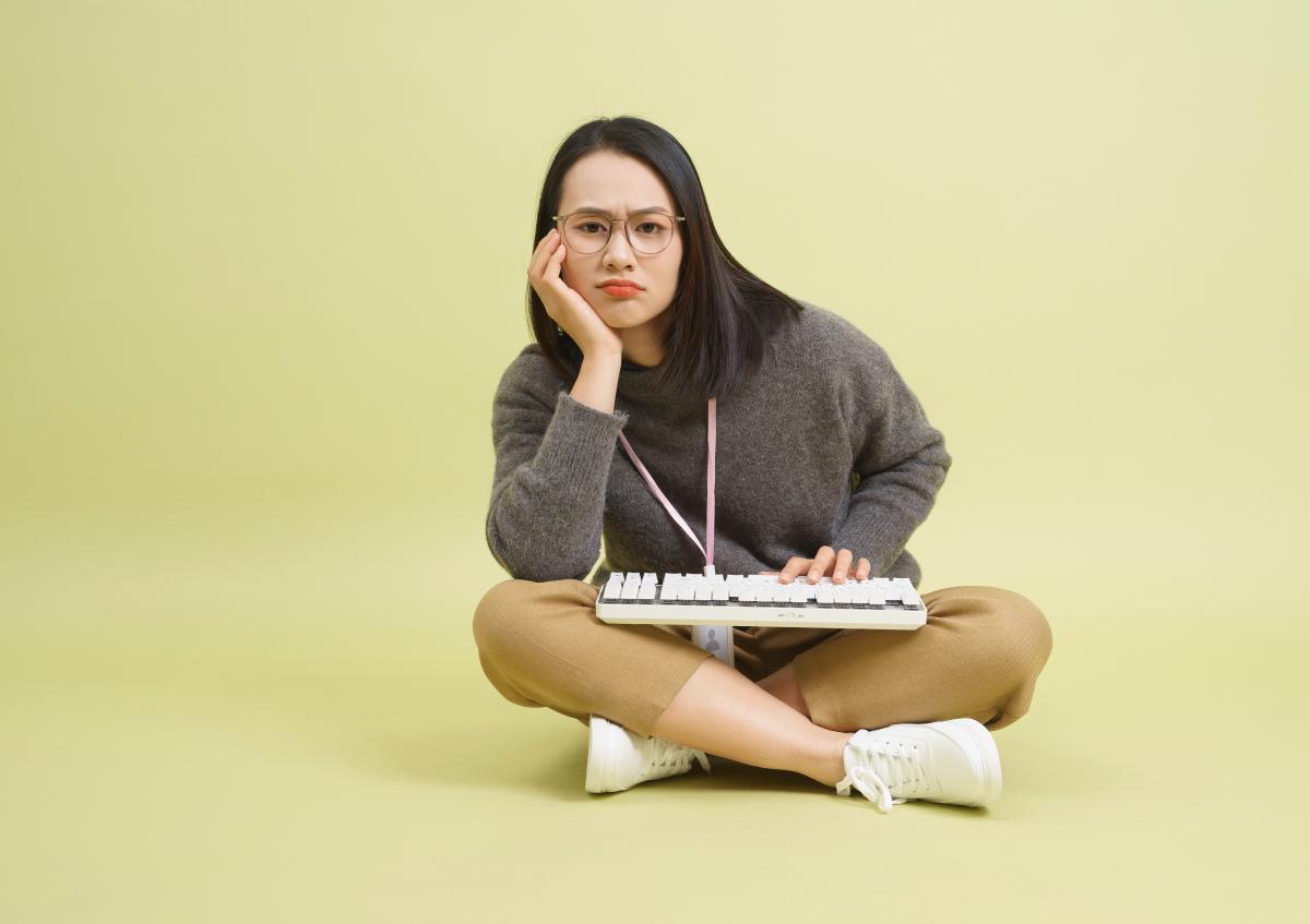 Vrouw zit met gekruiste benen op de grond, kijkt met een frons en licht beteuterde mond naar de camera. Ze ondersteunt haar hoofd met één hand en de andere hand rust op een toetsenbord dat op haar schoot ligt.