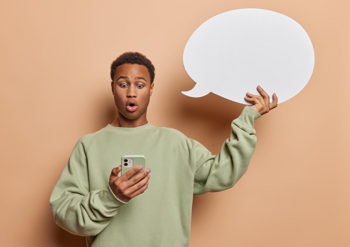 A young man holds a smartphone in one hand, looking at it with a surprised expression, mouth slightly open. With his other hand, he holds up a blank speech bubble.