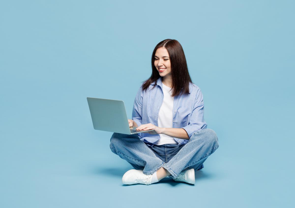 A woman sitting cross-legged on the floor, smiling while working on a laptop placed on her lap.