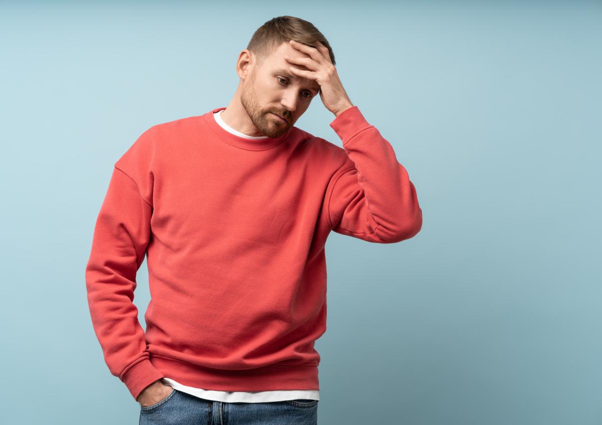 A man wearing a sweater looks worried or stressed as he holds one hand on his forehead and the other in his pocket.