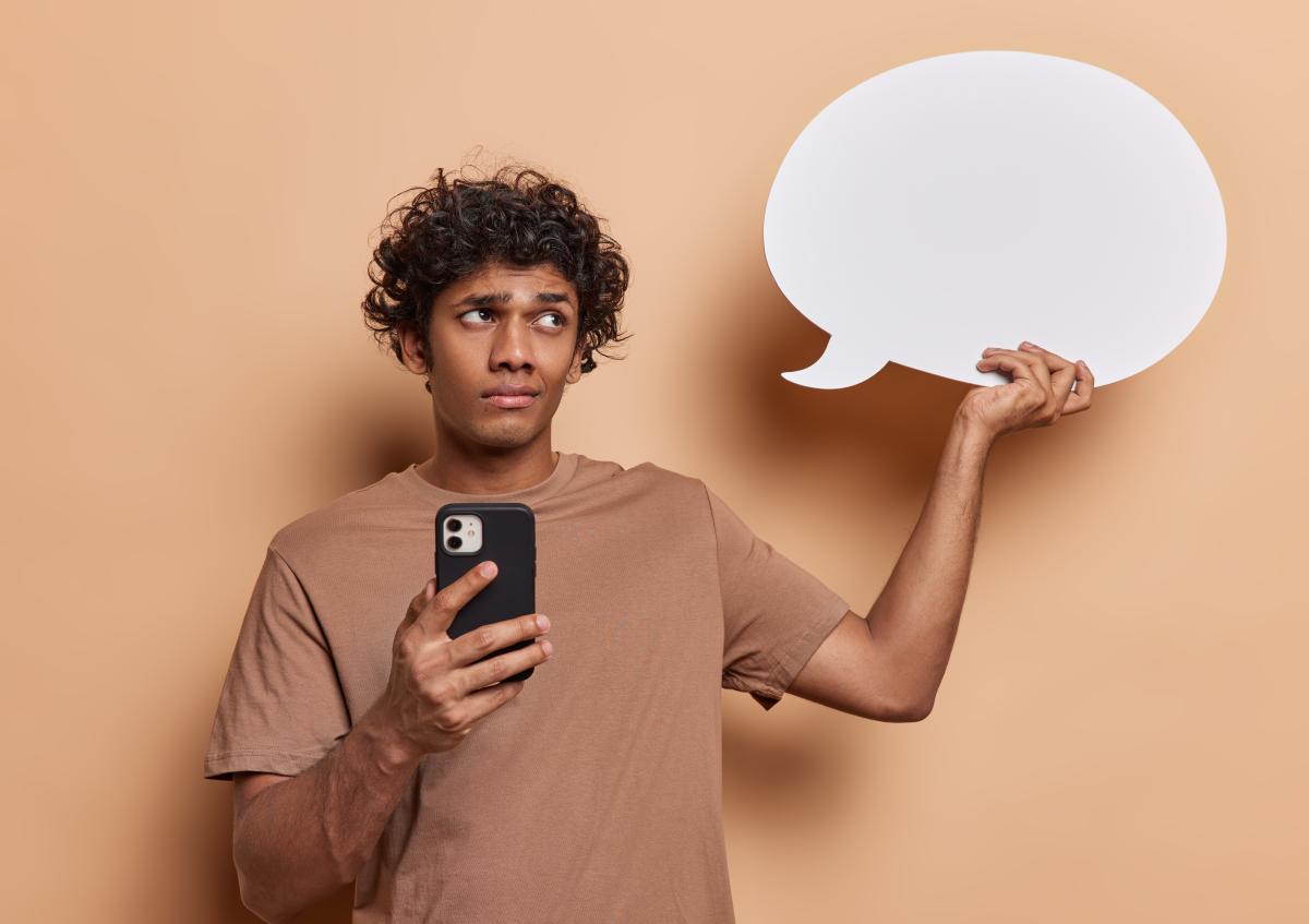 Een jonge man houdt met één hand een smartphone vast en met de andere een praatballonnetje. Hij kijkt opzij met een skeptische of twijfelende blik.