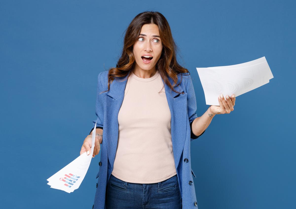 A surprised woman holds several sheets of paper in both hands, with her mouth slightly open and eyes wide, expressing shock or disbelief.