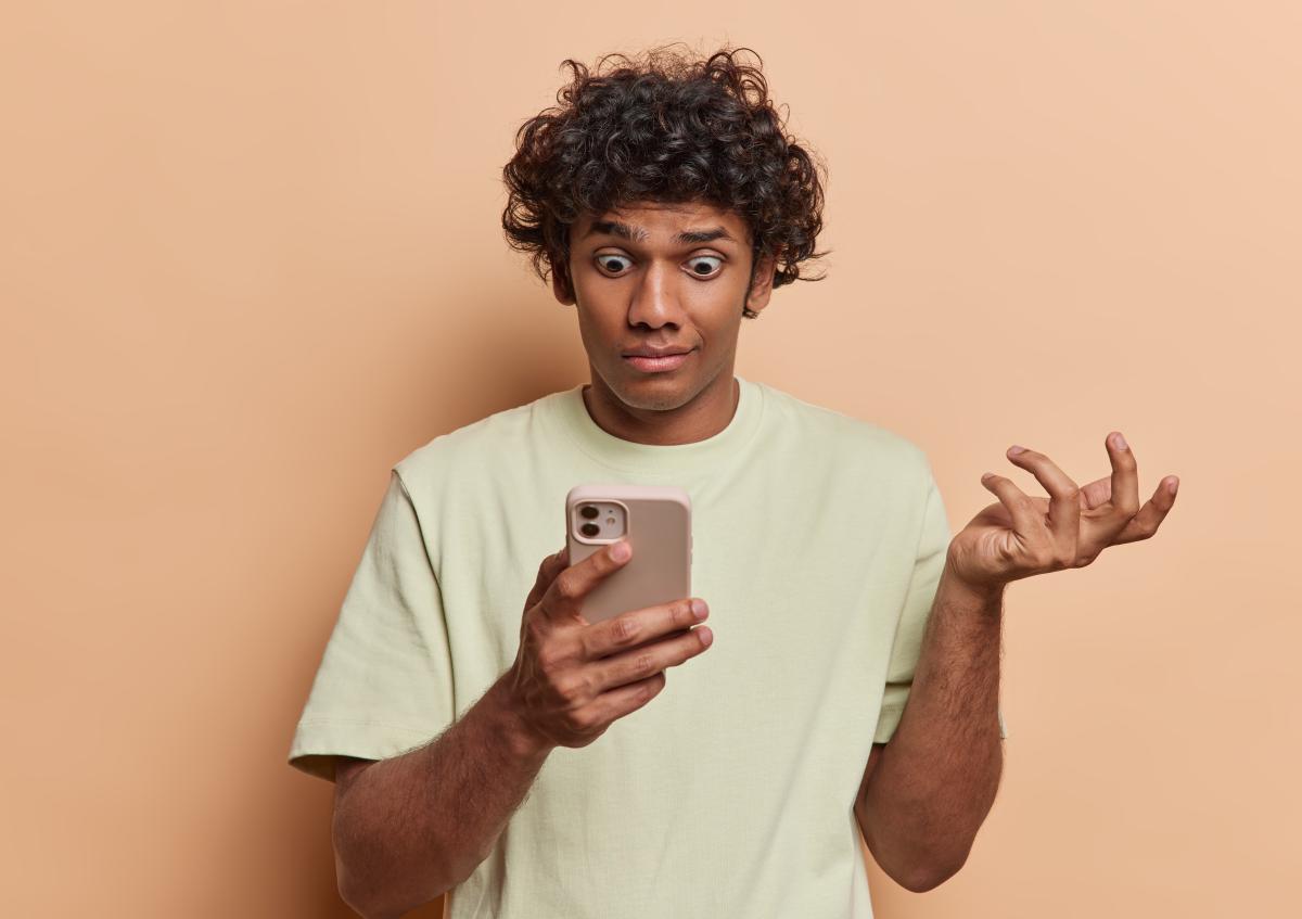 Een jonge man in een t-shirt kijkt verward en gefrustreerd naar zijn smartphone, terwijl hij met één hand omhoog gebaart uit verbazing.
