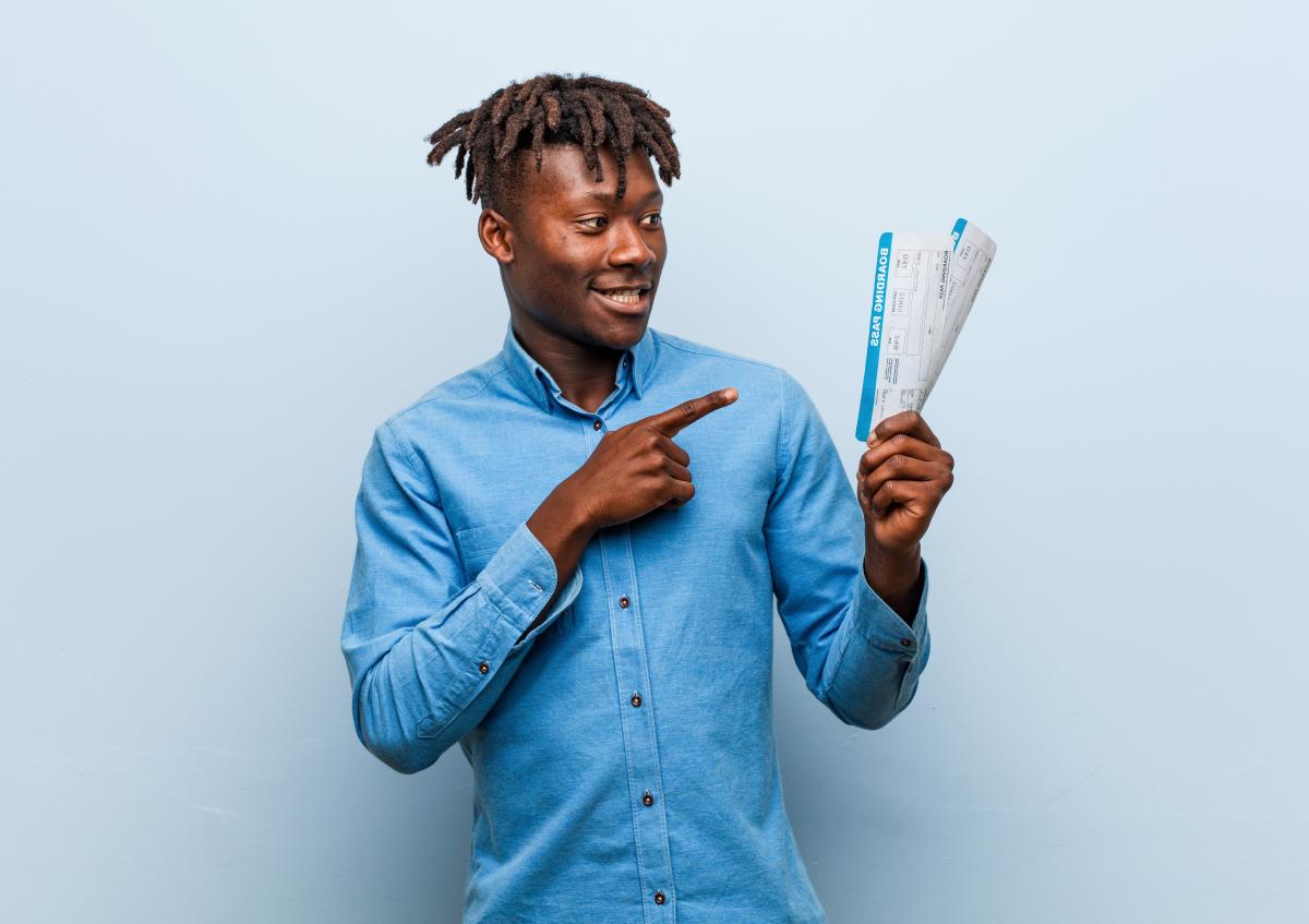 A young man smiling and pointing with his right hand to two boarding passes that he is holding in his left hand, suggesting excitement or readiness for travel