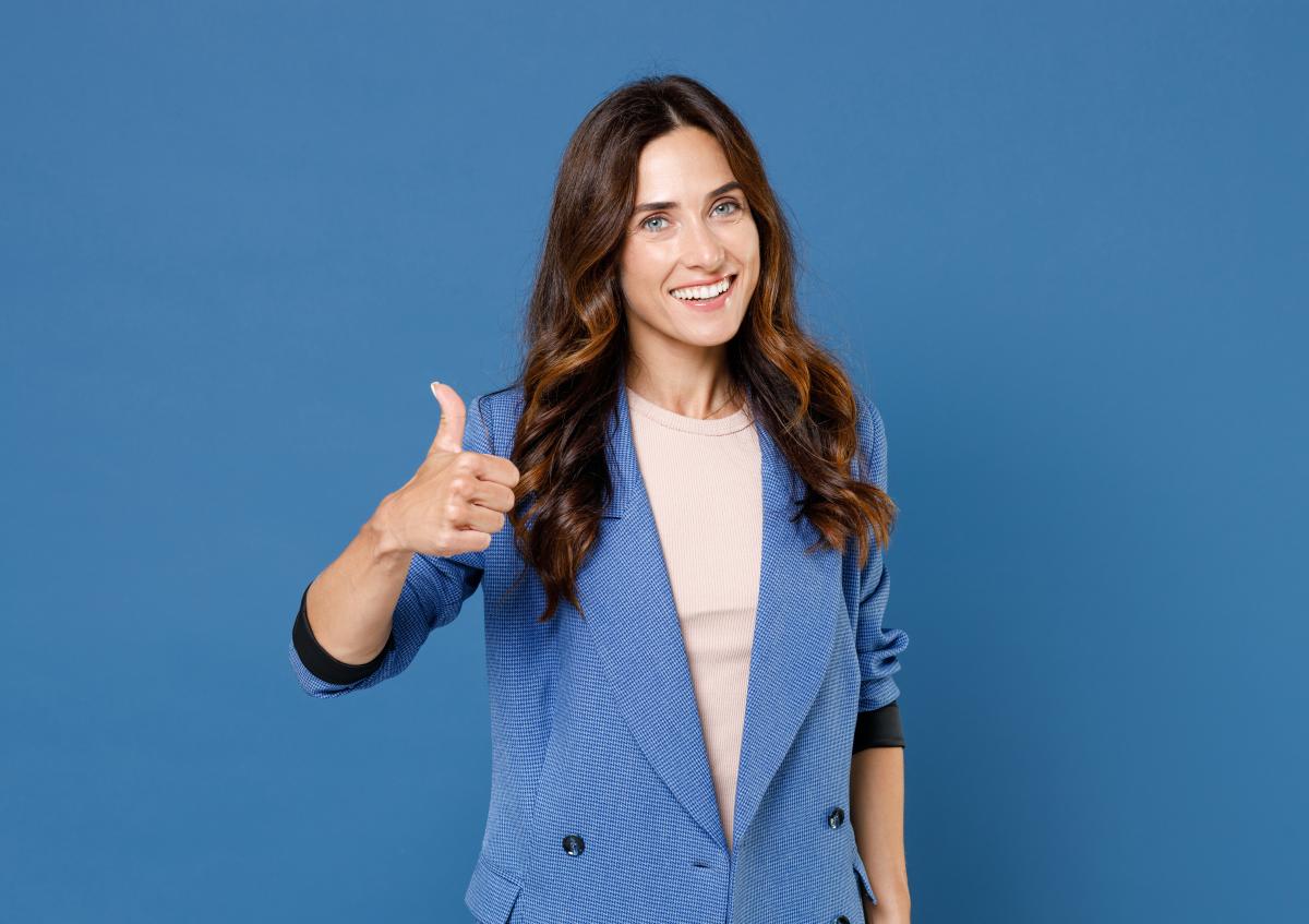 A confident woman smiling warmly, giving a thumbs-up with her right hand, showing approval or encouragement. 