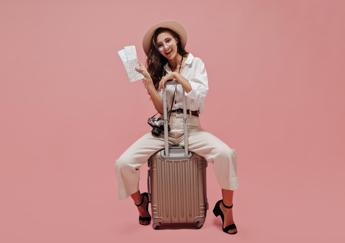 A cheerful young woman sitting on a suitcase, holding travel tickets in one hand and resting the other arm on the suitcase handle, smiling directly at the camera. She wears glasses and has a camera hanging around her neck.