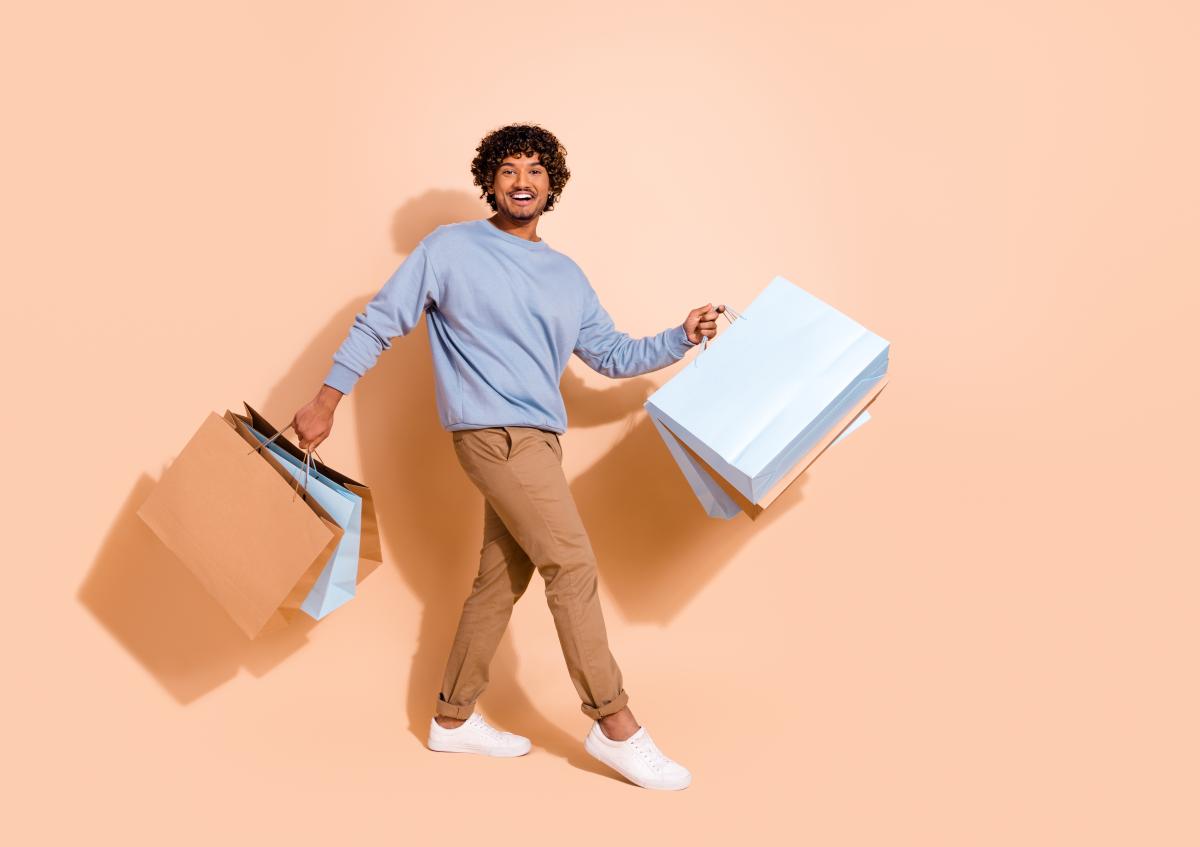 A man is walking while holding multiple shopping bags in both hands, smiling and looking joyful.