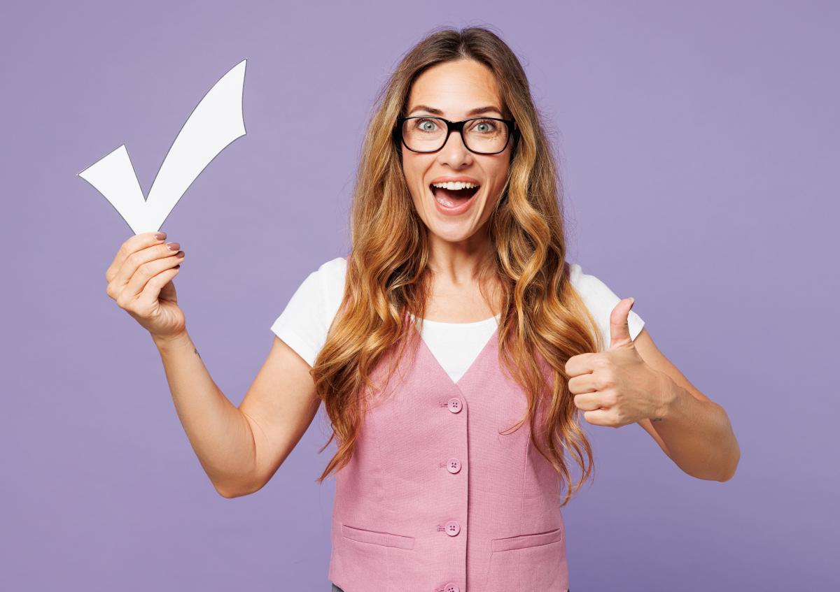 A woman with long hair and glasses smiles widely, holding a large paper checkmark in one hand and giving a thumbs-up with the other, expressing approval and positivity.