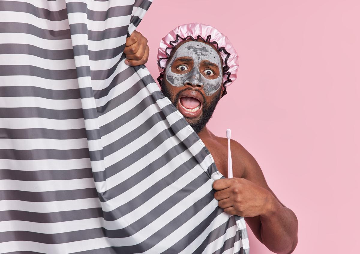 A man with a facial mask and shower cap peeks out from behind a striped shower curtain, holding a toothbrush and expressing surprise or shock with wide eyes and an open mouth.