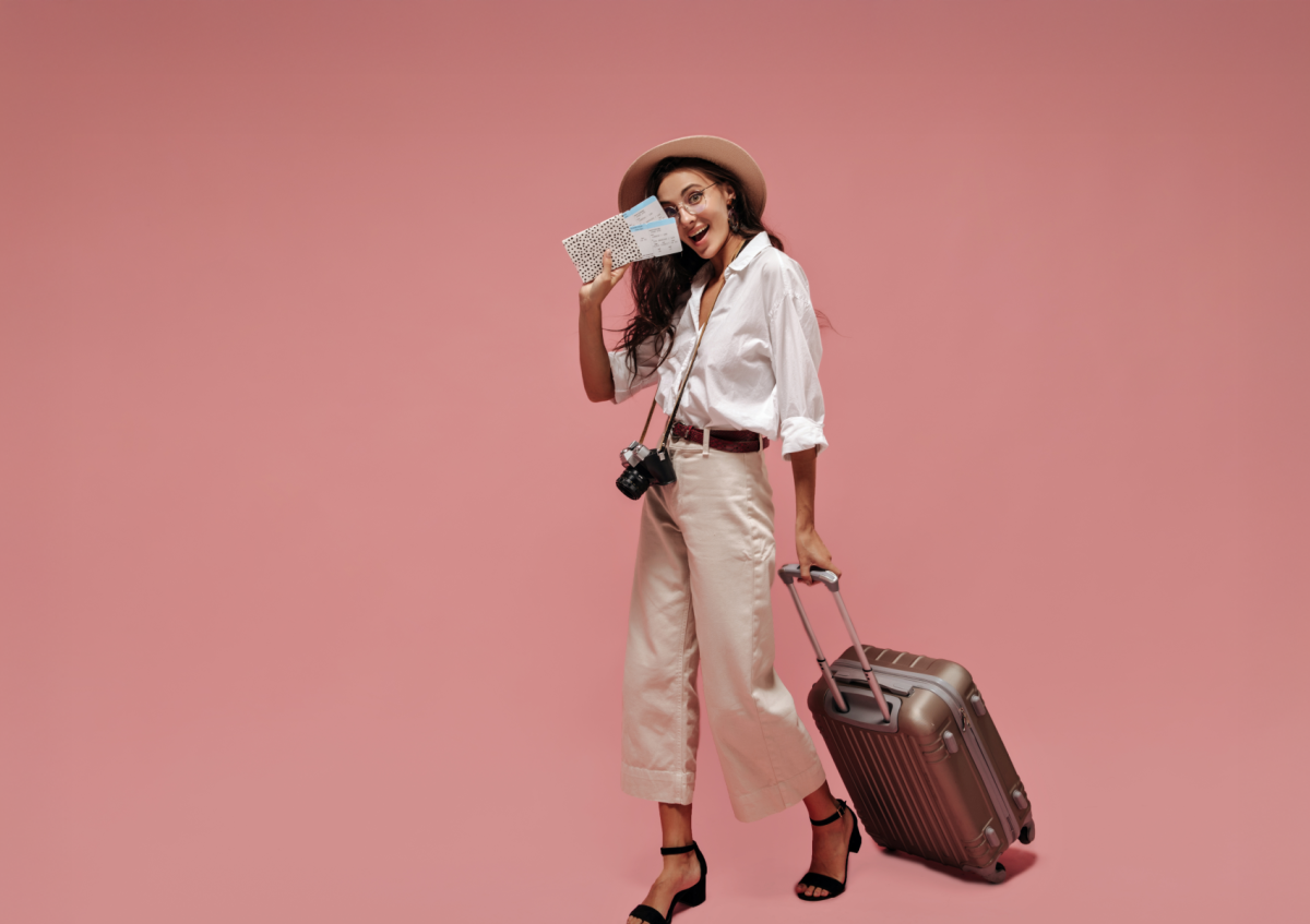 A smiling young woman in casual travel clothes holding boarding passes and a passport, pulling a suitcase, ready for a trip against a pink background