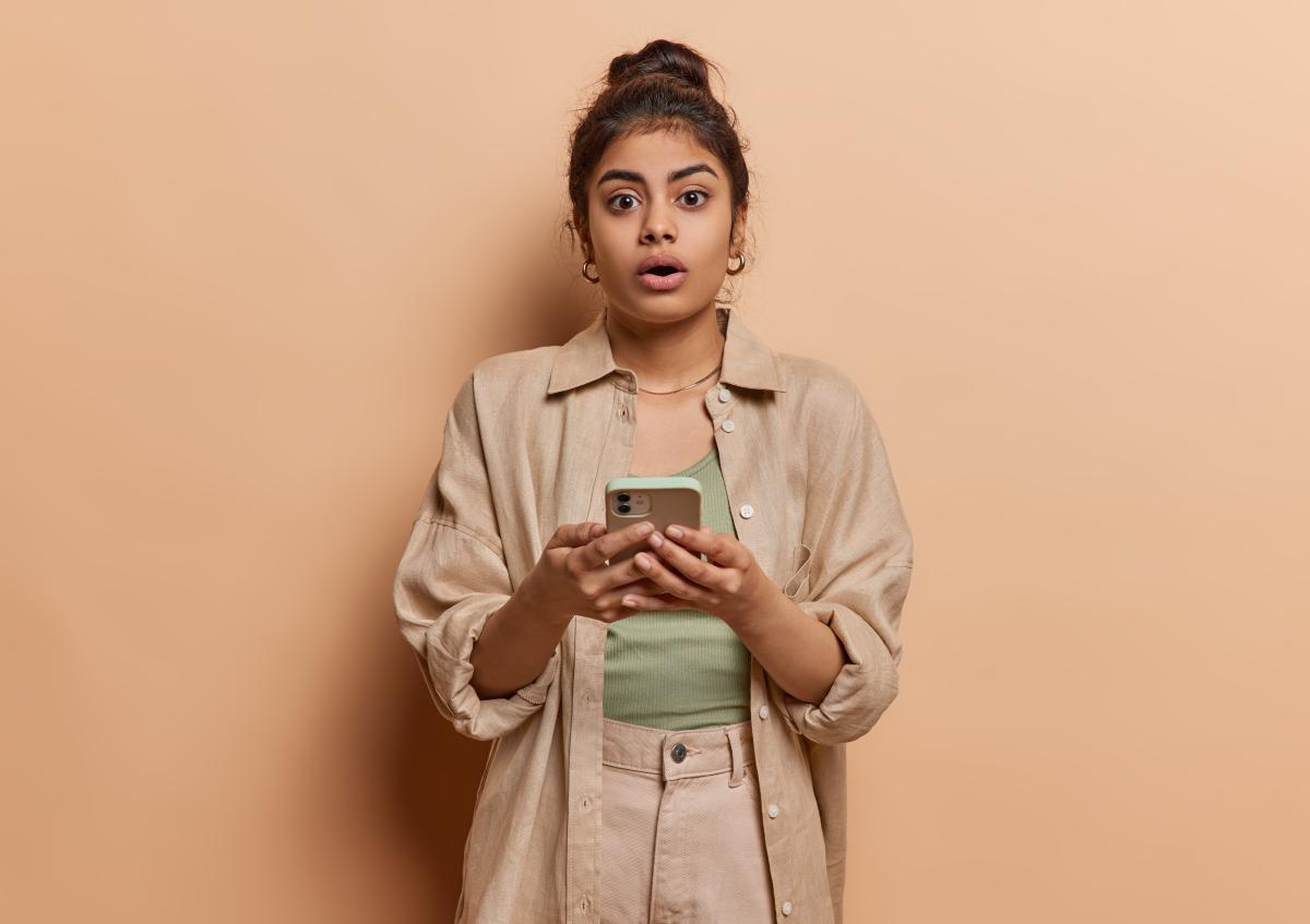 Person standing against a plain background, holding a smartphone with both hands and looking surprised or shocked, eyes wide and mouth slightly open.