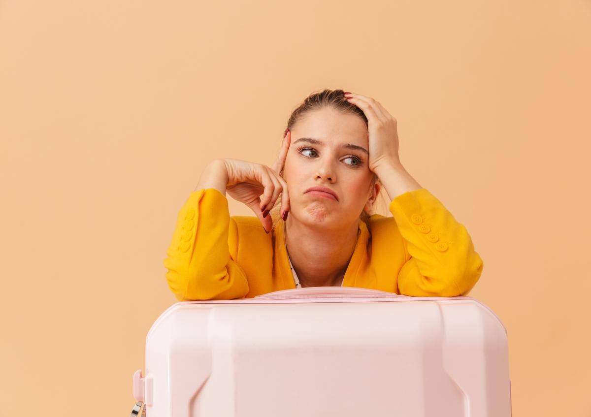 Woman with orange blazer leaning on pink suitcase, waiting for delayed flight. Peach-coloured background