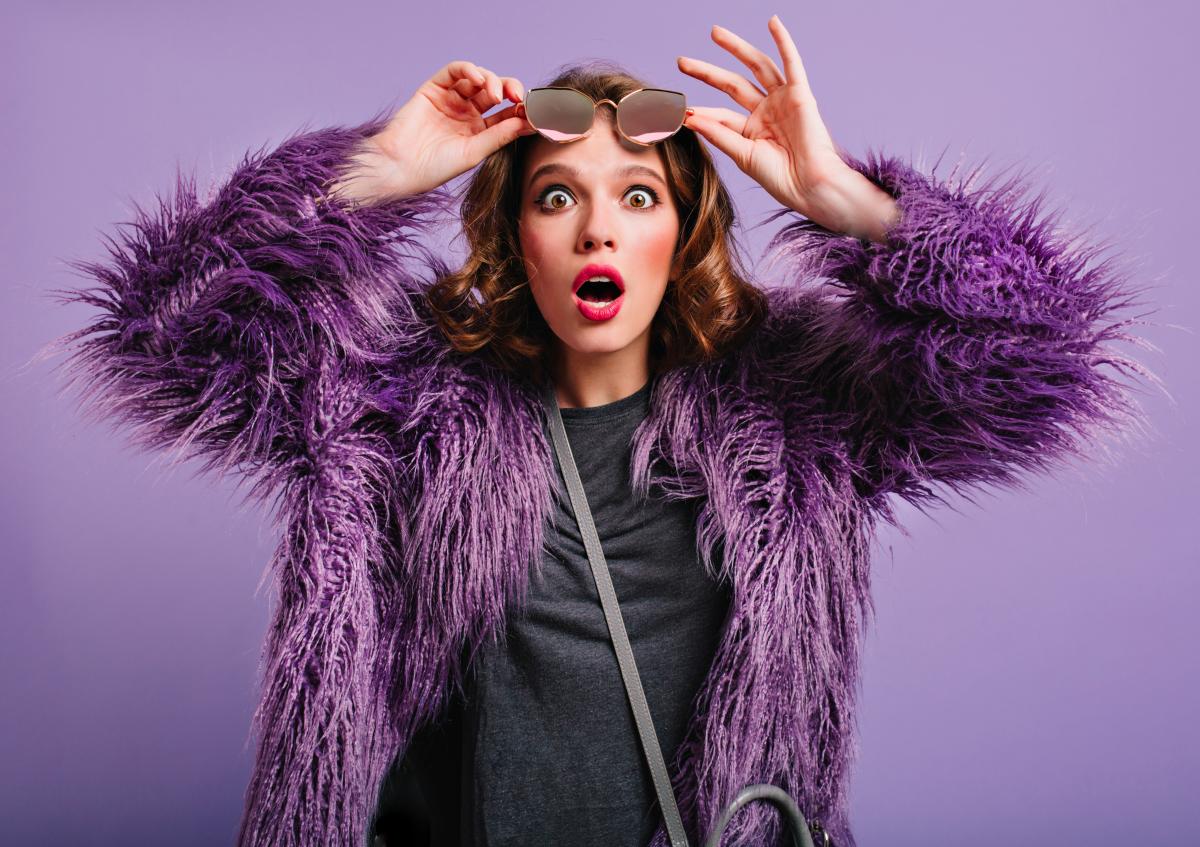 Vrouw met rode lipstick en paars jasje op paarse achtergrond kijkt verbaasd, houdt zonnebril omhoog