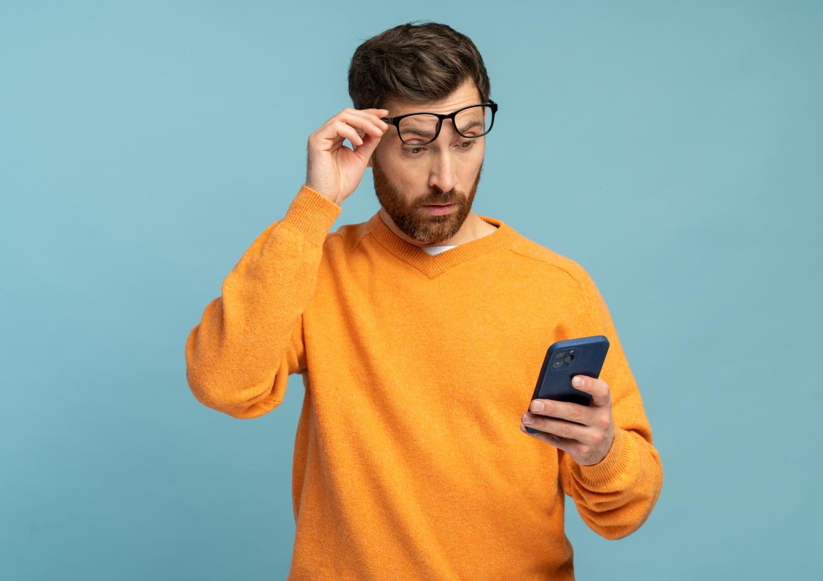 Man in orange jumper looks at phone and raises glasses when reading bankruptcy announcement - solid light blue background