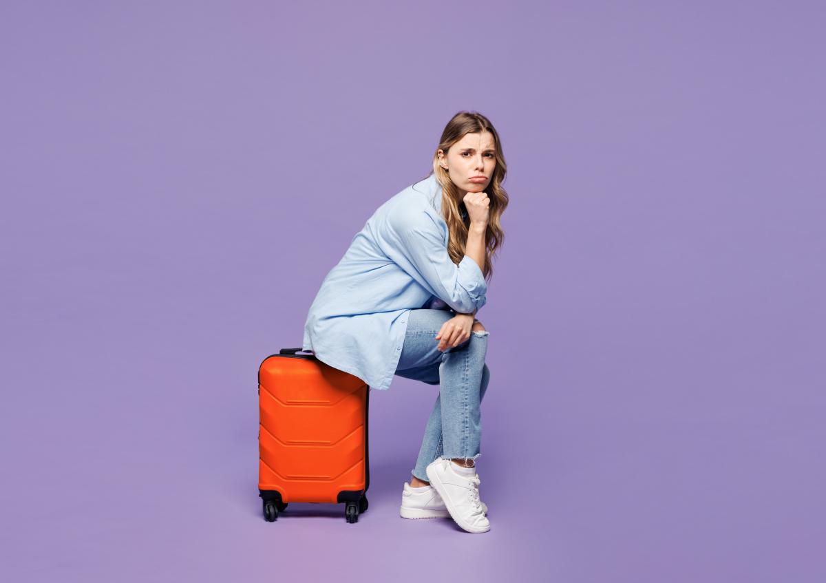 Woman in jeans and light blue blouse sits on red suitcase, waiting for her flight (uni purple background)