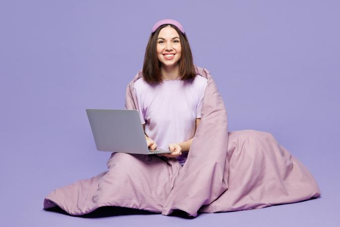 Young woman wrapped in a blanket, sitting with a laptop on her lap, smiling against, suggesting a cozy yet casual setting for streaming 