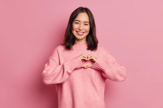 A smiling woman looking forward, making a heart shape with her hands in front of her chest.