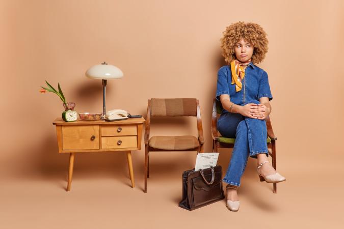 A young woman sitting on a chair next to an empty chair and a small table with a lamp, telephone, clock, and flower vase, looking away with an expression of frustration.