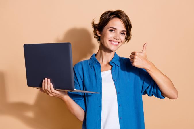Vrouw lacht vrolijk en kijkt tevreden in de camera. Ze houdt een laptop in haar linkerhand en steekt met haar rechterhand een duim omhoog.