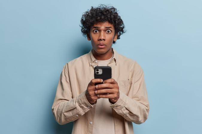 A young man holds a phone with both hands, looking at it with wide eyes and pursed lips, expressing surprise or shock.