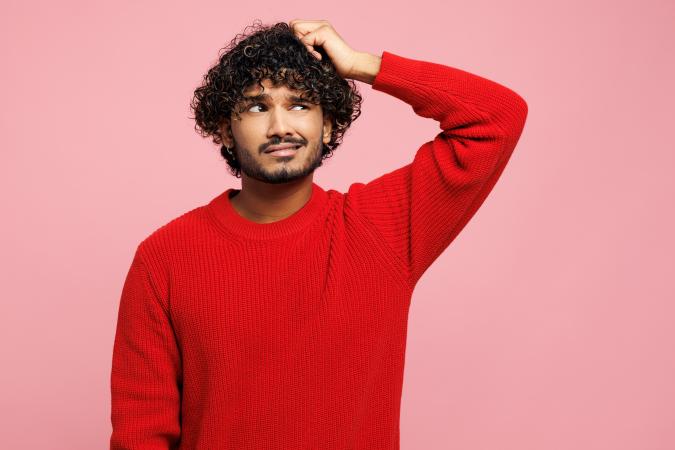 Een verwarde jonge man krabt met zijn linkerhand aan zijn hoofd en kijkt iets naar rechts met een vragende blik.