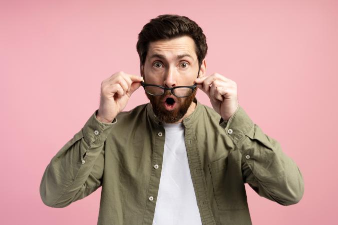 A man with a beard is holding his glasses down from his eyes, looking surprised and shocked, with his mouth open as if saying "wow" or expressing amazement.