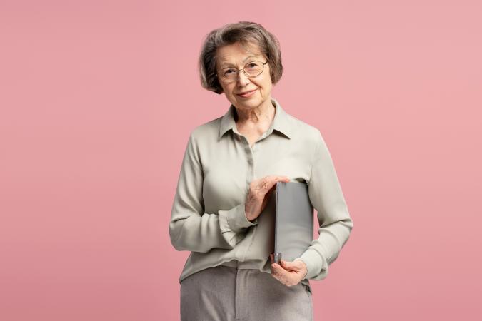 An elderly woman gently smiling, holding a closed laptop close to her chest with both hands.