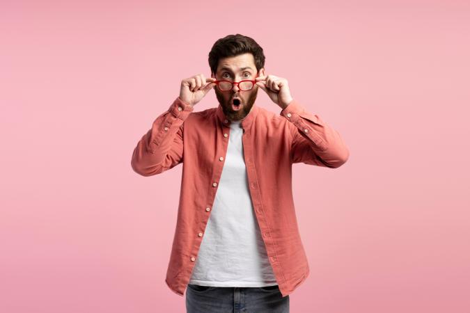 Man kijkt verbaasd door een rode bril, met open mond en opgetrokken wenkbrauwen, staand voor een roze achtergrond.