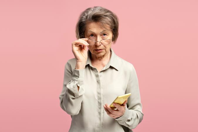 A serious older woman, holding a smartphone in one hand, and looking over the top of her glasses directly at the camera with a questioning expression