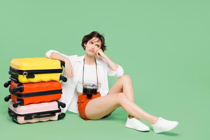 Vrouw zit op de grond met haar hand tegen haar wang, terwijl ze naast een stapel koffers zit. Ze kijkt omhoog met een frons en een lichte zucht, wat een uitdrukking van verveling of ongeduld toont.