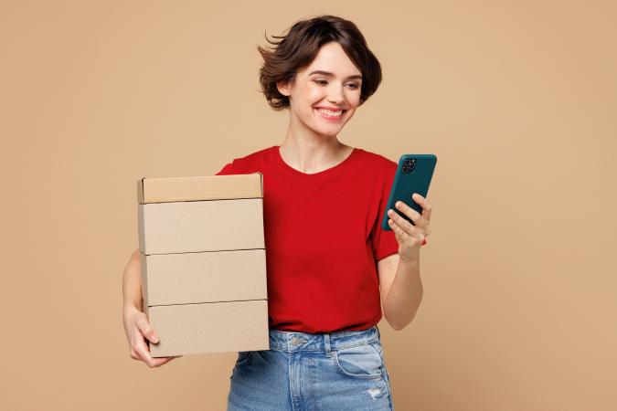 A smiling woman holding a stack of boxes in one arm and looking at a smartphone in the other hand.