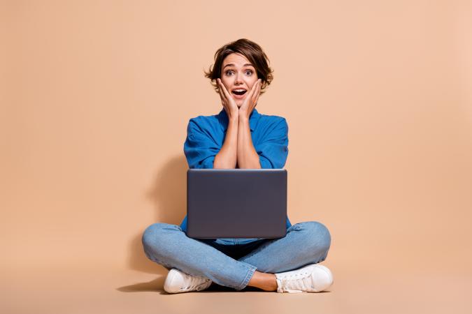 A shocked woman sits cross-legged on the floor with a laptop on her lap, holding her hands to her cheeks