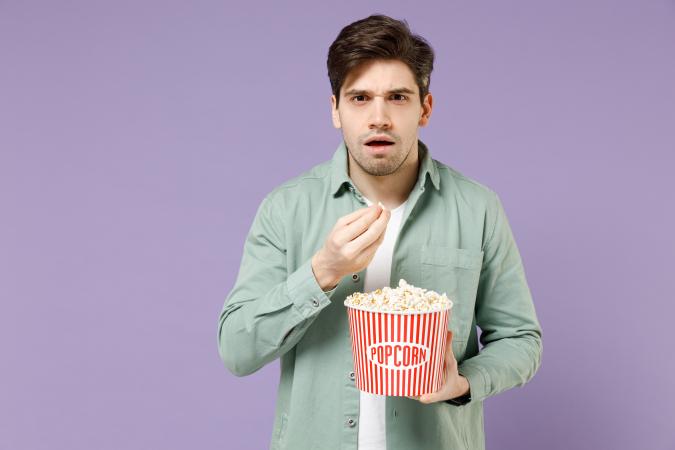 A young man holding a bucket of popcorn. His facial expression shows surprise or shock as he looks forward, holding a piece of popcorn near his mouth