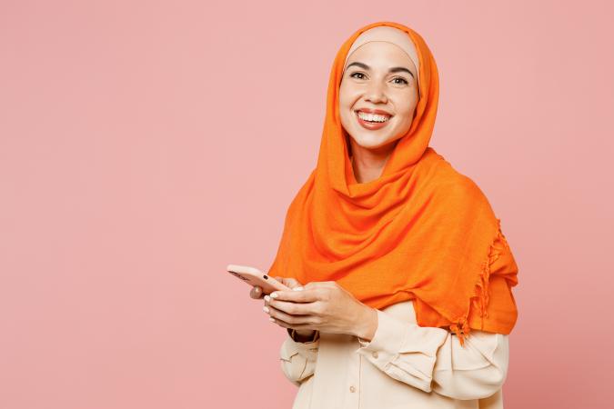 A smiling woman wearing a headscarf looks down at her phone while holding it with both hands, appearing happy and engaged.