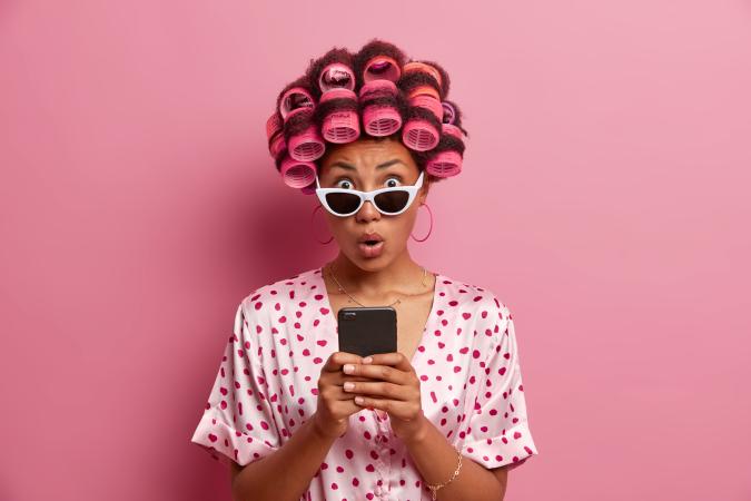 Vrouw kijkt met grote ogen en open mond verbaasd naar haar telefoon. Ze draagt een bril en heeft haar haar in krulspelden.