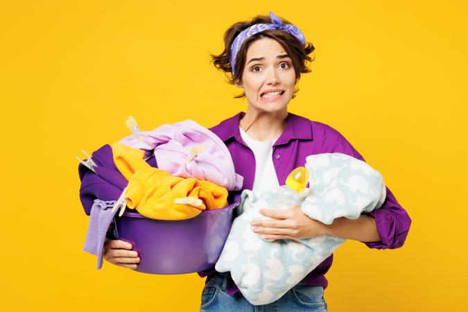A young woman holds a laundry basket filled with clothes. Her facial expression shows anxiety or stress as she bites her lip.
