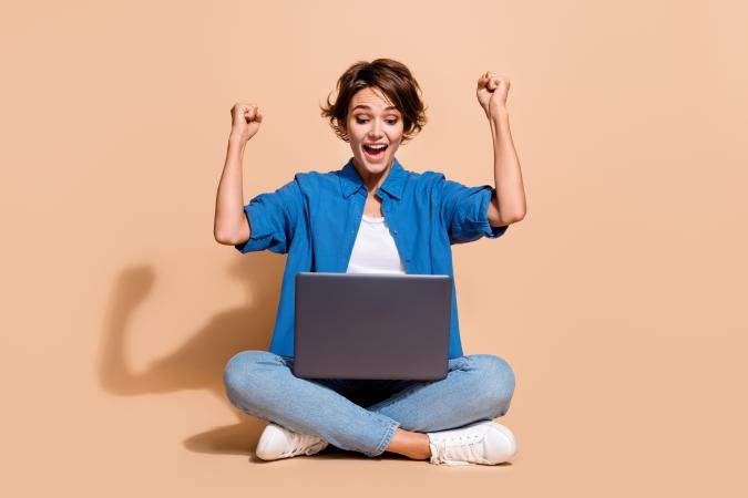 A young woman sits cross-legged on the floor, raising her arms in excitement while looking at a laptop screen.