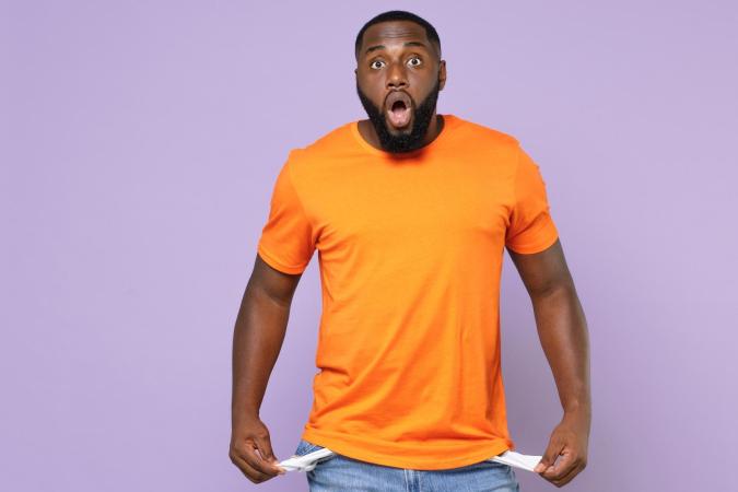 A surprised young man pulls out the empty pockets of his jeans, showing they contain no money. His mouth is open wide and his eyes are wide, expressing shock or disbelief