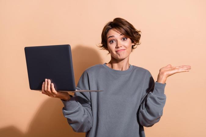 A young woman holding a laptop in one hand and raising her other hand with a shrug, wearing a confused or uncertain expression