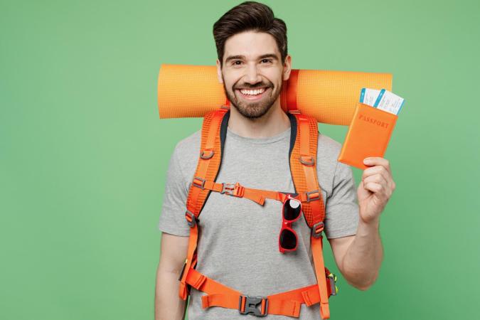 A young man with a beard, wearing a hiking backpack with a rolled-up sleeping mat, smiles broadly while holding a passport holder with boarding passes