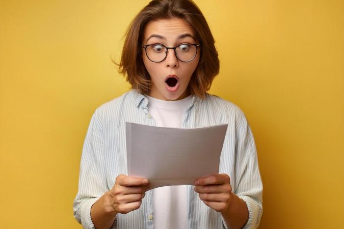 A surprised young woman holding a piece of paper in her hands and looks at it with wide eyes and an open mouth, expressing shock 