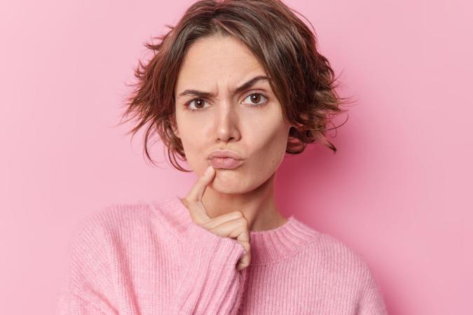 A young woman with an expression of confusion or puzzlement, with her eyebrows furrowed and her lips pursed. She is touching her chin lightly with her right index finger. 