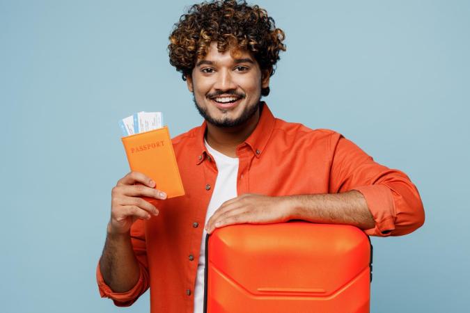 Man met gekruld haar en baard, glimlachend en een reiskoffer vasthoudend met zijn linkerarm. In zijn rechterhand houdt hij een paspoorthouder met vliegtickets erin.