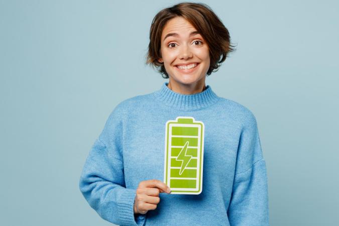 A young woman smiling widely and holding a large card with a fully charged battery symbol in front of her chest.