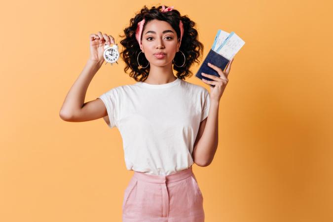 A young woman holding a small clock in one hand and a passport with boarding passes in the other, looking directly at the camera with a slightly pursed lip expression.