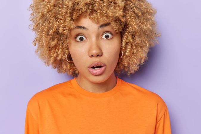 A young woman with curly hair wearing a bright top, looking directly at the camera with a surprised or shocked expression.