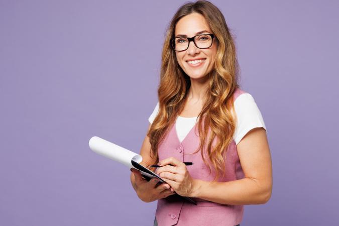 A woman smiling warmly while holding a notepad and pen, appearing ready to take notes or record information.