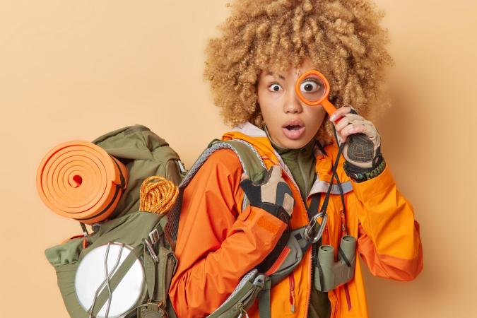 ChatGPT zei: A surprised woman dressed for outdoor adventure, carrying a backpack with camping gear and holding a magnifying glass up to one eye.